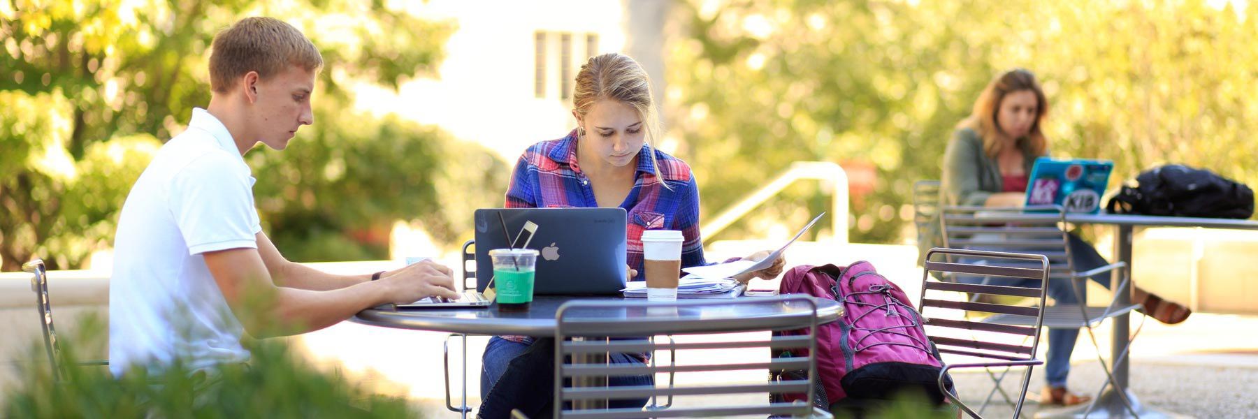 Students sit at a table outdoors and work on their laptops. 